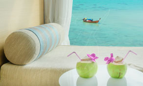Two young coconut drinks with pink straws and orchids on a white table beside a daybed overlooking turquoise sea and longtail boat