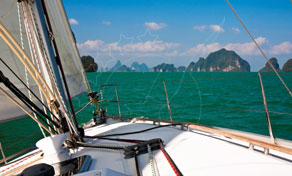 View from a sailboat bow across turquoise sea toward limestone karst islands under a hazy sky
