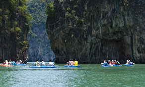 Tourists paddling canoes on green water beside towering limestone karst cliffs covered in jungle