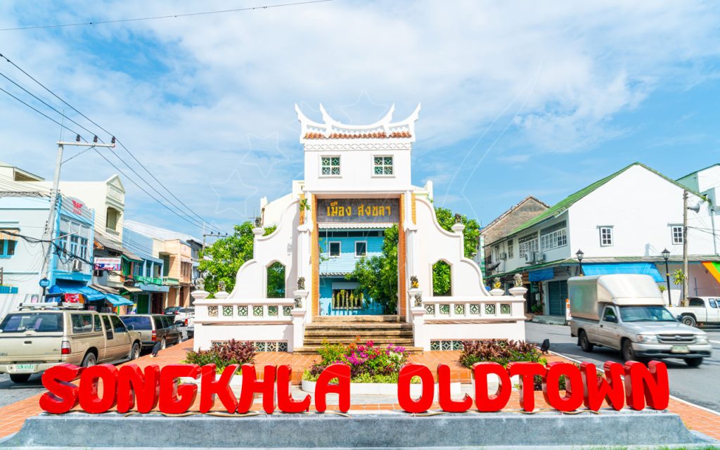 Red "SONGKHLA OLDTOWN" letters before a white Chinese-style gateway arch on a sunny Thai street