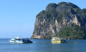 Two blue-and-white passenger ferries anchored in calm sea beside a jungle-covered limestone karst cliff under clear sky