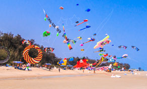 Colourful kites flying over a wide sandy beach under blue sky at a kite festival