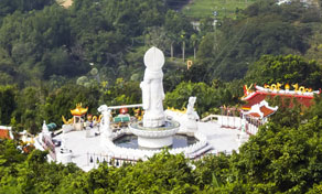 White Guanyin statue and shrine plaza with red-roofed pavilions on a green forested hilltop, seen from above