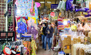 Shoppers walk a narrow indoor market aisle lined with stalls selling colourful clothing, bags and packaged snacks