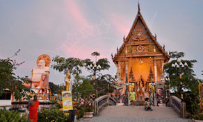 Thai Buddhist temple with golden gable and large seated Buddha statue at dusk, footbridge in foreground