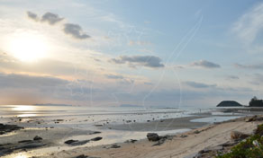 Sunset over a wide low-tide beach with wet sand, scattered rocks and a distant headland under hazy sky