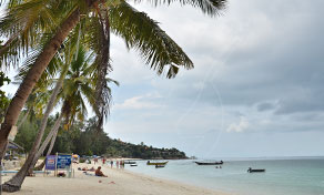 Palm-fringed white sand beach with longtail boats moored in turquoise shallows under an overcast sky