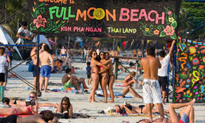 Crowded beach at Koh Phangan with a hand-painted "Welcome to Full Moon Beach" sign and people in swimwear on the sand