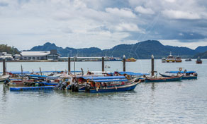 Traditional longtail boats moored at a pier with forested hills and overcast sky behind, Southeast Asia