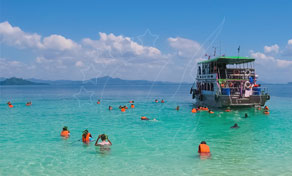 Snorkellers in orange life jackets swim in turquoise shallows beside a wooden tour boat near a tropical island