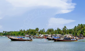Wooden longtail boats moored along a palm-fringed shoreline under a blue sky with scattered clouds in Thailand