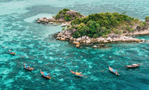 Aerial view of longtail boats moored in turquoise shallows beside a small rocky, tree-covered tropical island