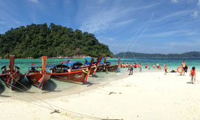Wooden longtail boats moored on a white sand beach with tourists and a green forested island under blue sky