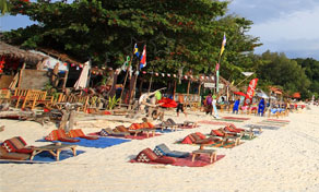 White sand beach lined with wooden sun loungers, colourful mats and flags outside rustic beachfront bars