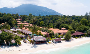 Aerial view of a beachfront resort with red-tiled roofs among palms, white sand beach and forested mountain