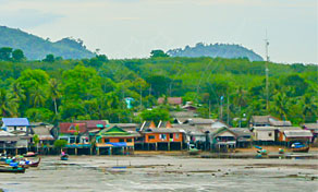 Stilt houses of a coastal fishing village at low tide, with longtail boats, mudflats and forested hills behind.
