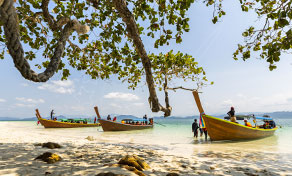 Longtail boats moored on a white sand beach with turquoise water, framed by overhanging tree branches in Thailand
