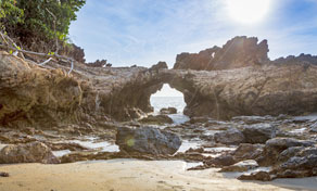 Natural rock arch on a secluded sandy beach with tide pools and sun flare over turquoise sea