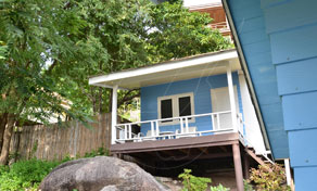 Blue wooden bungalow on stilts with a covered veranda, set above large boulders and tropical trees