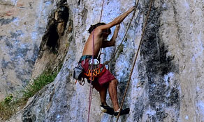 Shirtless climber in red shorts and harness scaling a grey limestone cliff face with ropes and quickdraws