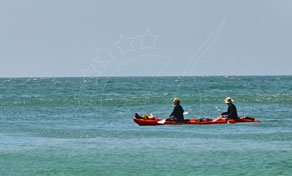 Two paddlers in a red sea kayak crossing calm turquoise water under a pale hazy sky