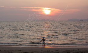Child standing at the water's edge on a sandy beach at sunset with a distant boat and islands on the horizon