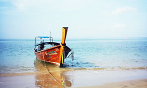 Wooden longtail boat with orange hull moored at the edge of a sandy beach beside calm turquoise water