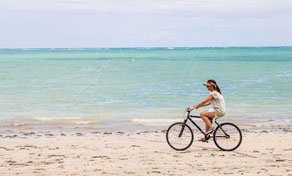Woman in white dress riding a black bicycle along wet sand beside a turquoise tropical sea