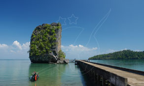 Limestone karst rising from calm turquoise sea beside a long concrete pier with a moored longtail boat under blue sky