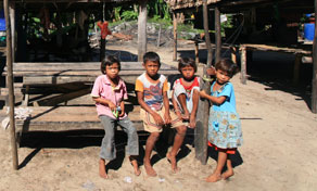 Four young children sit on a wooden bench on sandy ground in front of a stilt house in a rural tropical village