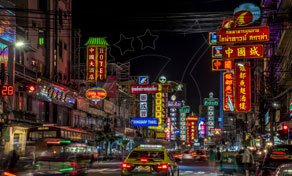 Neon signs in Thai and Chinese light up a busy Bangkok Chinatown street at night with cars and taxis in traffic