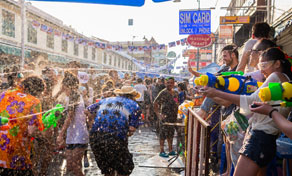 Crowds spraying water guns during Songkran street festival on a Bangkok road lined with shops and a SIM card sign