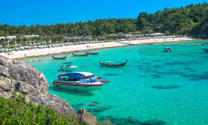 Longtail and speed boats moored in turquoise bay beside white sand beach backed by forested hills