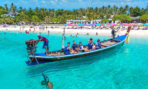 Longtail boat with passengers in turquoise shallow water off a busy tropical beach lined with palm trees