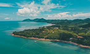 Aerial view of a forested tropical headland with a jetty, turquoise sea and green hills under cloudy blue sky