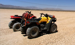 Two parked quad bikes, one yellow and one red, on cracked dry desert flats with hazy mountains behind