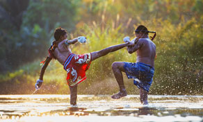Two Muay Thai fighters sparring in shallow water at golden hour, one landing a high kick amid splashes