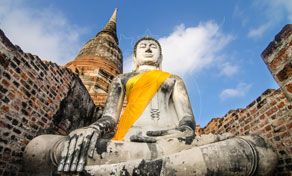 Seated stone Buddha statue draped in orange sash amid brick temple ruins with a golden chedi under blue sky