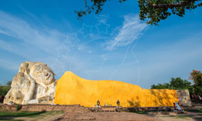 Large reclining Buddha statue draped in bright yellow cloth under a clear blue sky, framed by tree branches
