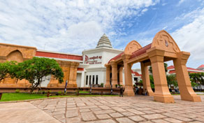 Ornate arched gateway of red and cream masonry framing a white Thai museum building across a paved plaza under blue sky