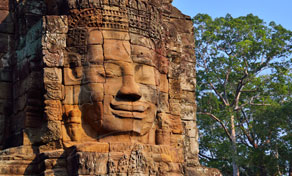 Weathered stone face carved into a sandstone tower at Bayon temple, Angkor, framed by green trees and blue sky