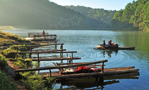 Bamboo rafts moored along a misty lake shore with people paddling a raft past forested hills at sunrise