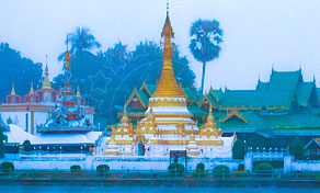 Golden Burmese-style temple with tiered spire beside a lake at dusk, palm trees and hills behind