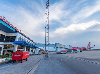 Red AirAsia jet parked at a regional Thai airport terminal with jet bridge, floodlight mast and ground equipment under blue sky