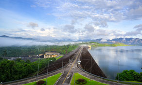 Road along a large dam crest beside a calm reservoir, with misty limestone mountains and green hills at sunrise