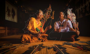 Two musicians seated on a woven mat play traditional stringed instruments in a dimly lit room