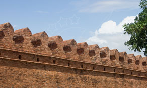 Ancient red brick city wall with crenellated battlements under a blue sky, green tree leaves at right