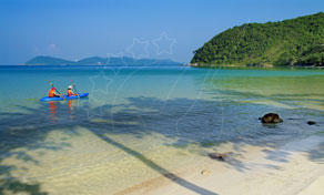 Two people paddle a blue kayak in shallow turquoise water off a sandy beach with a forested headland behind
