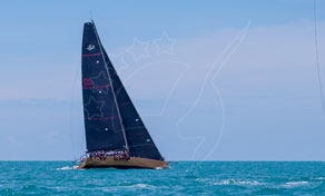 Dark-hulled racing yacht under full sail heeling on calm open sea beneath a hazy blue sky