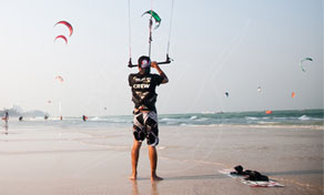 Kitesurfer standing on a sandy beach holding a control bar, with many kites flying over the sea behind